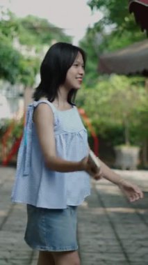 Yavaş çekim genç Asyalı kız bahçenin yanındaki bir barın terasında elinde akıllı telefonuyla el sallıyor ve gülümsüyor.