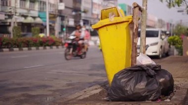 Tayland 'da plastik poşetlerle çevrili büyük sarı bir çöp tenekesinin yanında yavaş hareket eden araba ve motosiklet trafiği - 4K yatay video