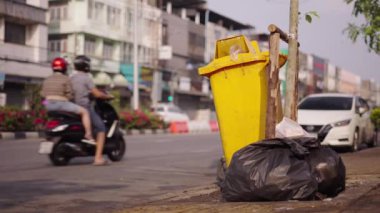 Bangkok 'ta işlek bir caddede, büyük sarı bir çöp kutusunun yanında yanlış yönde giden bir motosiklet. 4K yatay video.