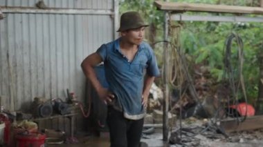 Genç, kirli ve ıslak Asyalı tamirci Vietnam 'daki küçük aile kırsal atölyesinde sigara içiyor - 4K Yatay