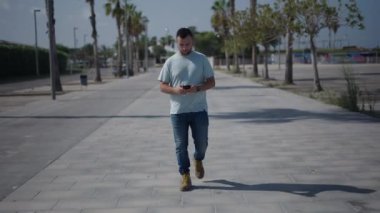 Palmiye ağaçları arasındaki beton geçitte yürüyen beyaz çocuk akıllı telefonundan aldığı bir mesaja cevap veriyor. 4K yatay video.
