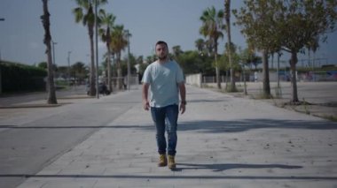 Kafkasyalı genç kameraya doğru yürüyor cebinde akıllı telefonuyla palmiye ağaçları arasındaki beton yol boyunca el sallıyor - 4K yatay video