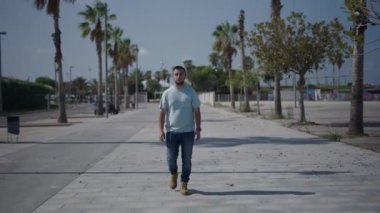 Beyaz çocuk kameraya doğru yürüyor cebinde akıllı telefonuyla palmiye ağaçları arasındaki beton yolda - 4K yatay video