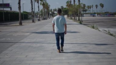 Palmiye ağaçları arasındaki çimento geçidi boyunca kameraya doğru yürüyen beyaz çocuk, akıllı telefonundan aldığı bir mesaja cevap veriyor. 4K yatay video.