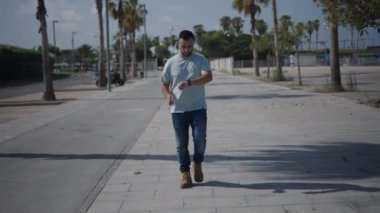 Palmiye ağaçları arasındaki çimento geçidi boyunca kameraya doğru yürüyen beyaz bir çocuk akıllı sesiyle bir ses notu gönderiyor - 4K Yatay Video