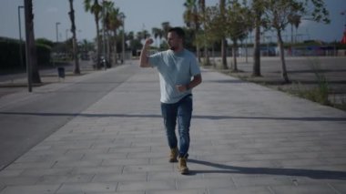 Palmiye ağaçları arasındaki beton geçitte kameraya doğru yürüyen beyaz çocuk favori takımını alkışlıyor ve alkışlıyor. 4K Yatay Video.