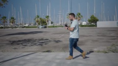 Palmiye ağaçları arasındaki beton yolda yürüyen beyaz bir çocuk profil olarak plastik bardaktan kahvesini yudumlayarak 4K Yatay Video 'ya geçiyor.