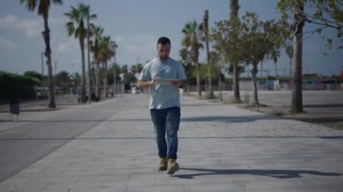 Palmiye ağaçları arasındaki çimento geçidi boyunca kameraya doğru yürüyen beyaz bir çocuk akıllı sesiyle bir not gönderiyor - 4K Yatay Video