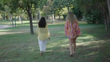 Bir esmer ve bir sarışın, sırtı kameraya dönük çimenlerin üzerinde yalın ayak yürüyüp sohbet ediyorlar.