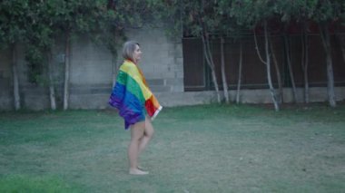 Beyaz, genç bir kız çimenlerde çıplak ayakla yürüyor kameradan yüzünü çeviriyor arkasında bir LGBT bayrağıyla gülümsüyor - 4K Yatay