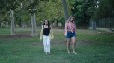 İki genç beyaz kız bir esmer ve bir sarışın çimde yalın ayak yürüyorlar kameraya doğru gülümsüyorlar - 4K Yatay