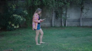 Genç, sarışın, beyaz bir kız, profilden yürüyor. Çimlerin üzerinde kameraya doğru yürüyor. Tabletin dokunmatik pedi üzerinde yazıyor.
