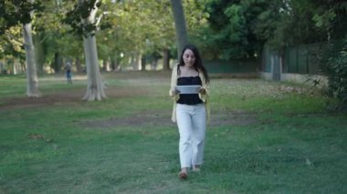 Genç esmer beyaz kız çimenlerin üzerinde kameraya doğru yürüyor gülümsüyor elinde tabletle bir video görüşmesi yapıyor - 4K Yatay