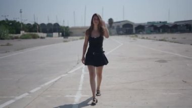 Beyaz, kısa siyah elbiseli genç bir kız limanın yakınındaki sanayi bölgesinde kameraya doğru yürüyor ve baş parmakları aşağıda, 4K yatay hareket yapıyor.