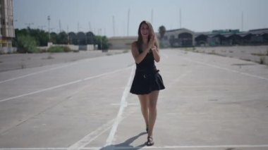 Beyaz, kısa siyah elbiseli genç bir kız limanın yakınındaki sanayi bölgesinde kameraya doğru yürüyor. El çırpıyor. 4K Yatay.