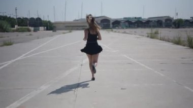 Beyaz kısa siyah elbiseli genç bir kız sırtı kameraya dönük bir şekilde limanın yakınındaki sanayi bölgesinde yürüyor - 4K Yatay