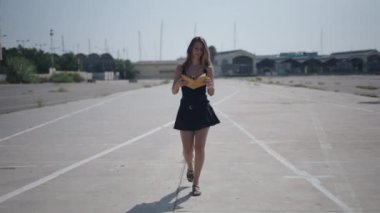 Beyaz kısa siyah elbiseli beyaz bir kız liman yakınlarındaki banliyö sanayi bölgesinde kameraya doğru yürüyor ve kitap okuyor - 4K Yatay