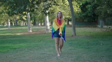 Arkasında LGBT bayrağıyla çimenlerin üzerinde çıplak ayakla yürüyen genç beyaz kız. 4K Yatay Bayrak.