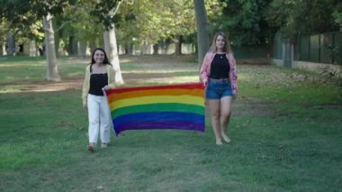 Bir esmer ve bir sarışın, sırtlarında bir LGBT bayrağıyla kameraya dönük olarak çimlerin üzerinde çıplak ayakla yürüyorlar.
