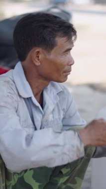 Asyalı olgun bir adam çok kirli bir tişörtle çömelmiş Vietnam 'da taşınabilir bir soğutucudan su içiyor.