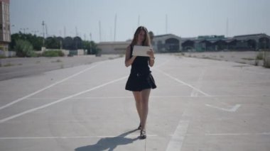 Beyaz kısa siyah elbiseli genç bir kız limanın yakınındaki sanayi bölgesinde kameraya doğru yürüyor ve tabletinin ekranıyla etkileşime geçiyor - 4K Yatay