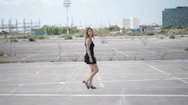 Kısa siyah elbiseli beyaz bir kadın profilden yürüyor ve limanın yakınındaki sanayi bölgesinde kameraya öpücük yolluyor. 4K Yatay.