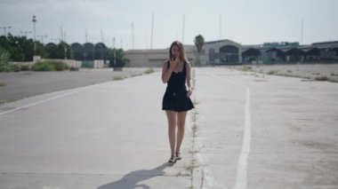 Beyaz, kısa siyah elbiseli genç bir kadın limanın yakınındaki sanayi bölgesinde kameraya doğru yürüyor. Akıllı telefonuyla bir sesli mesaj gönderiyor. 4K Yatay.