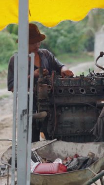 Ağır çekimde genç bir tamirci tehlikeli bir ortamda zayıf güvenlikli eski bir traktörün motorunu tamir ediyor.