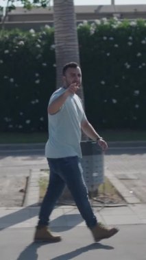 Kafkasyalı genç, palmiye ağaçları arasındaki beton yürüyüş yolunda kameraya doğru yürüyor ve FullHD dikey video görüntülerini gösteriyor.