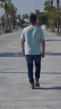 Palmiye ağaçları arasındaki çimento geçidi boyunca kameraya doğru yürüyen beyaz çocuk, akıllı telefonundan aldığı mesaja cevap veriyor.