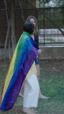 Arkalarında LGBT bayrağı, FullHD dikey bayrağıyla çimenlerin üzerinde sırtüstü yürüyen bir sarışın ve bir esmer genç beyaz kız.