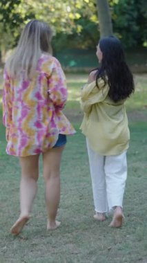 İki beyaz kız, bir esmer ve bir sarışın, kameraya danışmak ve akıllı telefonlarını paylaşmak için sırtüstü çimenlerin üzerinde çıplak ayakla yürüyorlar.