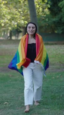 Beyaz, genç, sarışın bir kız kameraya bakmadan çimenlerde yalın ayak yürüyor arkasında bir LGBT bayrağıyla gülümsüyor.
