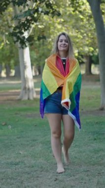 Beyaz, genç bir kız, çıplak ayakla çimlerin üzerinde yürüyor kameradan yüzünü çeviriyor arkasında bir LGBT bayrağı var FullHD dikey