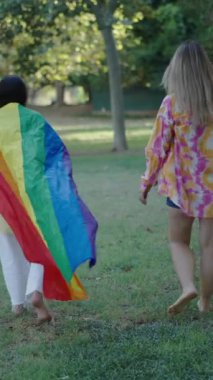 Bir esmer ve bir sarışın çıplak ayakla çimlerin üzerinde yürüyorlar kameraya dönük bir LGBT bayrağıyla arkalarında FullHD dikey