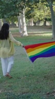 Bir esmer ve bir sarışın çıplak ayakla çimlerin üzerinde yürüyorlar kameradan uzağa bakıyorlar aralarında bir LGBT bayrağı tutuyorlar- FullHD Dikey