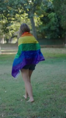 Bir esmer ve bir sarışın, kameraya doğru çıplak ayakla yürüyorlar ve aralarında bir LGBT bayrağı sallıyorlar.
