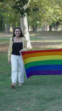 Bir esmer ve bir sarışın, sırtlarında bir LGBT bayrağıyla kameraya dönük olarak çimlerin üzerinde yalın ayak yürüyorlar.