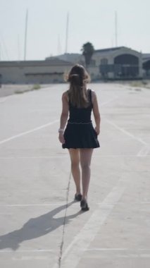 Kısa siyah elbiseli genç beyaz kadın, sırtı kameraya dönük bir şekilde limanın dışındaki sanayi bölgesinde yürüyor.