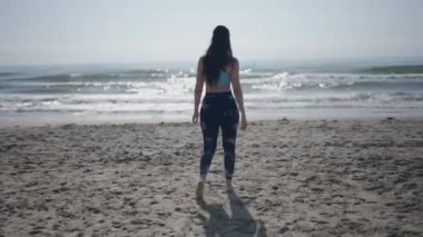 Genç, uzun saçlı, beyaz saçlı, at kuyruklu bir kadın güneşli bir günde kumsalda sırtı kameraya dönük yürüyor - 4K Yatay