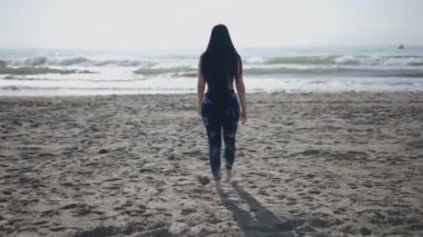 Uzun saçlı, esmer, beyaz bir kadın güneşli bir günde kumsalda sırtı kameraya dönük yürüyor ve kameraya el sallıyor - 4K Yatay