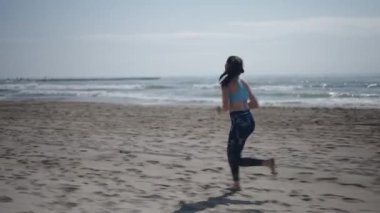 Kumsaldaki çıplak ayaklı kumsalda uzun esmer saçlı ve spor yapan genç bir kız. 4K yatay.