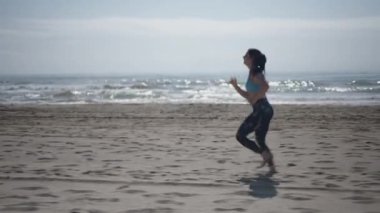 Kumsalın kumsalında çıplak ayakla koşan ve profilden gülen uzun esmer saçlı beyaz bir kadın.