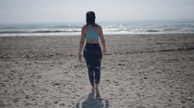 Genç, uzun saçlı esmer, beyaz bir kadın güneşli bir günde kumsalda geriye doğru yürüyen ellerini hareket ettirerek hayranları - 4K Yatay