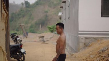 Genç Vietnamlı şortlu ve çıplak gövdeli adam sigara içtikten sonra ağzından duman üflüyor - 4K Yatay