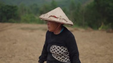 Yorgun, yetişkin bir Vietnamlı kadın çiftçinin portresi iş günü kafasında büyük bir konik şapkayla - 4K Yatay