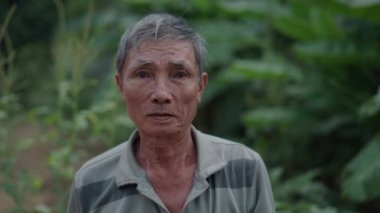 Nehir ve doğa yakınlarındaki kırsal bir alanda kameraya gülümseyen yaşlı bir Vietnamlı erkek çiftçinin portresi - 4K Yatay