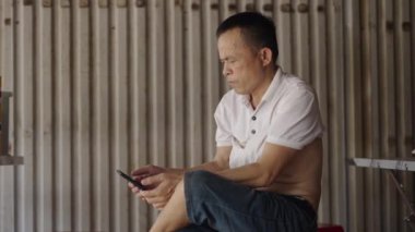 Sandalyede oturan olgun bir Asyalı Vietnam 'daki akıllı telefon ekranından haberleri okuyor - 4K Yatay