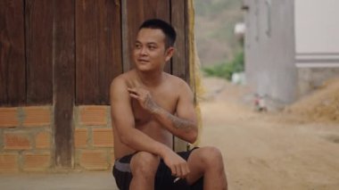Çıplak gövdeli genç Vietnamlı bir adam elinde sigarasıyla evinin kapısında oturuyor. 4K Yatay.