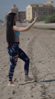 Genç, uzun saçlı esmer, beyaz kadın güneşli bir günde kumsalın kumsalında profilden yürüyen akıllı telefonuyla selfie çekiyor.